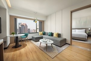 A modern hotel room with a large window, a sofa, a dining table, and a bed.
