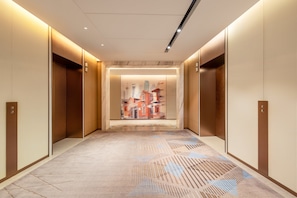 Un couloir avec des ascenseurs, une moquette Ă motifs et une Ćuvre dâart reprĂ©sentant un paysage urbain accrochĂ©e au mur.