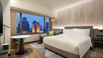 A hotel room with a large bed, a small table, a chair, a TV, and a view of a cityscape at dusk.
