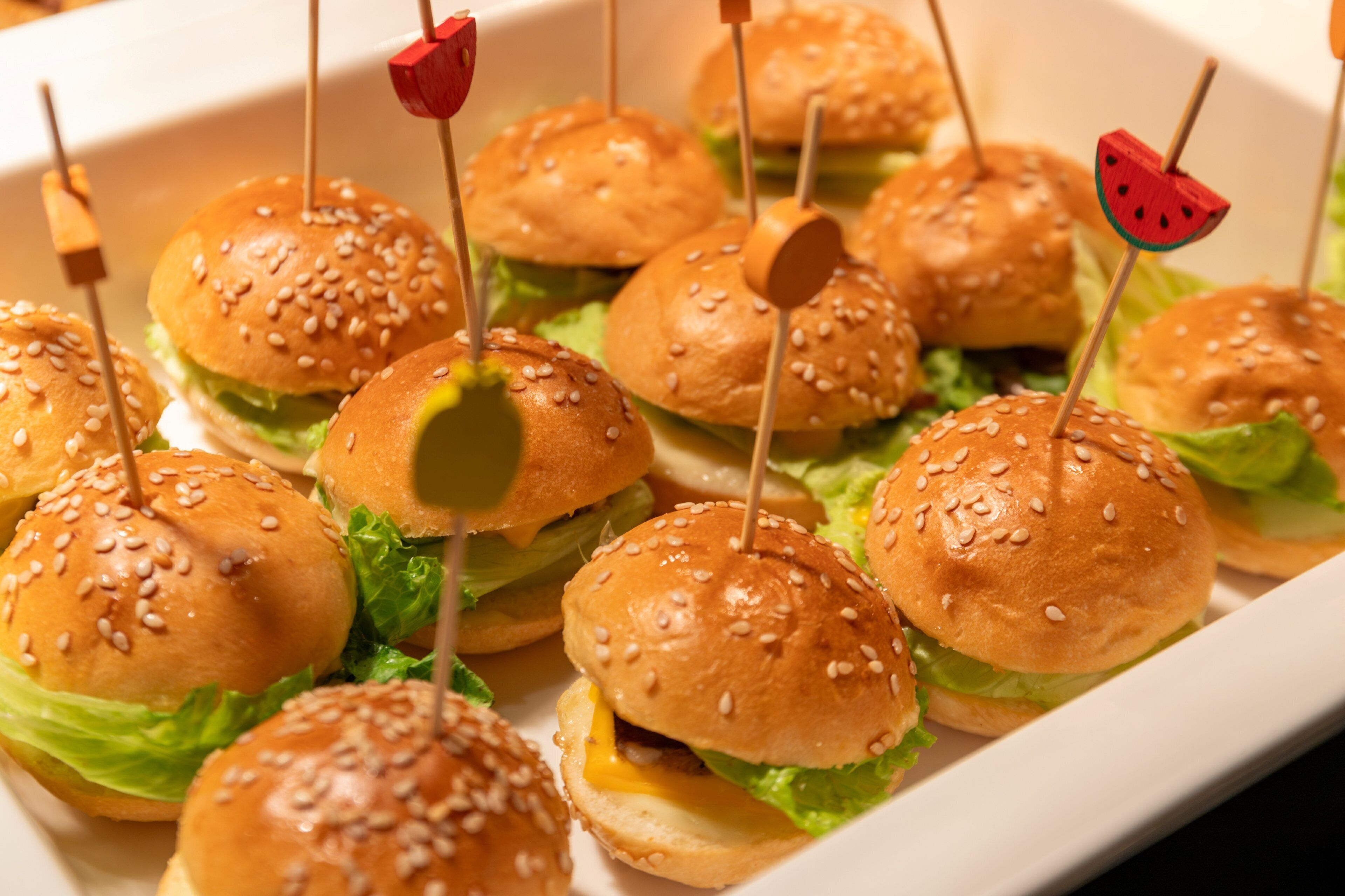 A tray of mini sandwiches with sesame seed buns and toothpicks.
