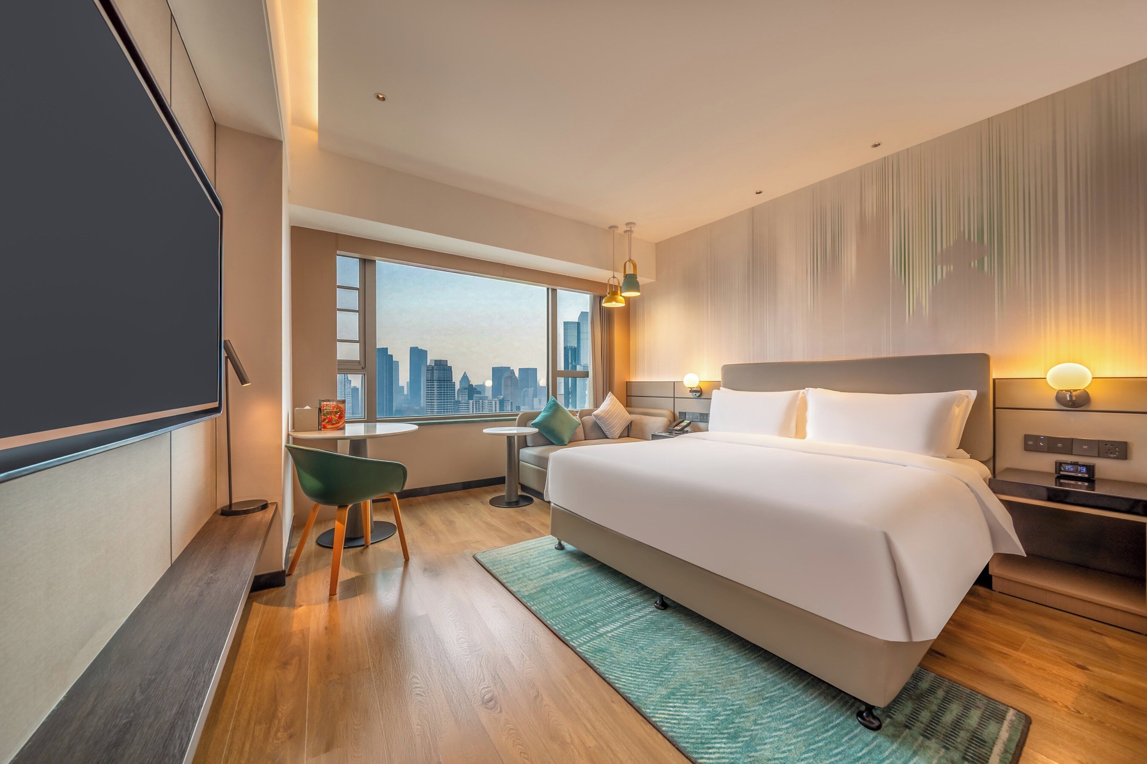 A hotel room with a large bed, a desk, a chair, a view of the cityscape, and a city skyline visible through the window.