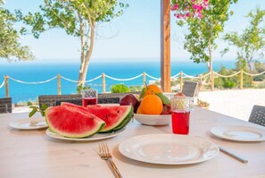 A table set with plates, cutlery, a bowl of fruit, and a glass of red drink, overlooking a beach and the sea. - Villa Paradiso Sunset (Pomos)
