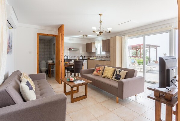 A modern living room with a sofa, coffee table, and a television. There is a kitchen area with cabinets and a dining table. - Villa Paradiso Sunset (Pomos)