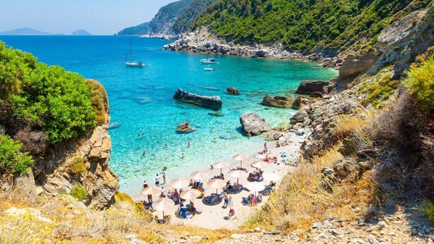 Ein Küstenstrand mit klarem türkisblauem Wasser, felsigem Gelände und üppiger Vegetation.
