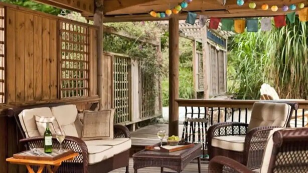 A covered outdoor seating area with wicker furniture, a wooden table, and a bottle of wine. - Plan Your Escape to the Cottage! Minutes to Mystic, Connecticut. This pet-friendly retreat has a private fenced-in rear garden patio. Your stay includes organic breakfast, firepit, hammock, and onsite hiking trails. Up to 2 well-behaved pets welcome! (Ledyard)