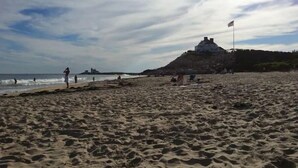 A beach with a castle-like structure on a hill, people walking, and a flag on a pole. - Plan Your Escape to the Cottage! Minutes to Mystic, Connecticut. This pet-friendly retreat has a private fenced-in rear garden patio. Your stay includes organic breakfast, firepit, hammock, and onsite hiking trails. Up to 2 well-behaved pets welcome! (Ledyard)