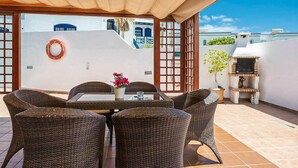 A covered outdoor dining area with wicker furniture, a table with a vase of flowers, and a barbecue grill.