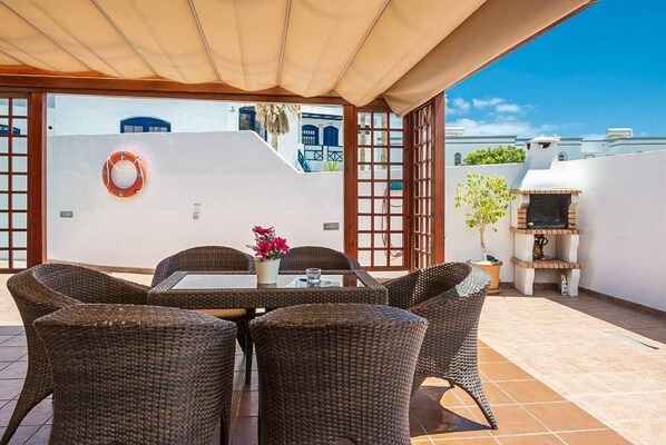 A covered outdoor dining area with wicker furniture, a table with a vase of flowers, and a barbecue grill.