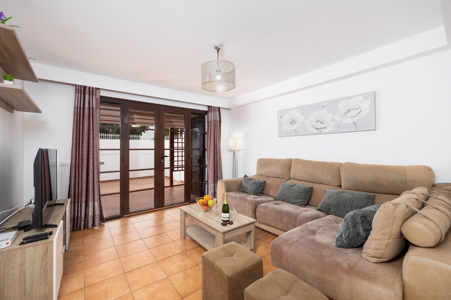 A living room with a sofa, a coffee table with fruit and a bottle, a TV, and a sliding glass door leading to an outdoor area.
