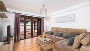 A living room with a sofa, a coffee table with fruit and a bottle, a TV, and a sliding glass door leading to an outdoor area.