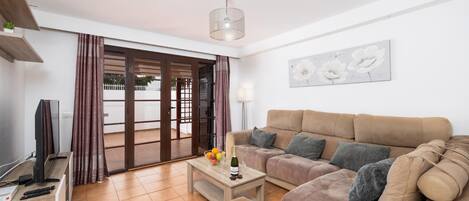 A living room with a sofa, a coffee table with fruit and a bottle, a TV, and a sliding glass door leading to an outdoor area.