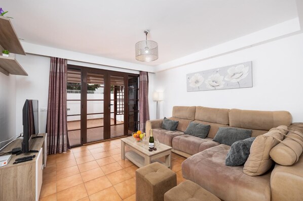 A living room with a sofa, a coffee table with fruit and a bottle, a TV, and a sliding glass door leading to an outdoor area.
