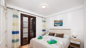 A hotel room with a bed, a nightstand, a fan, and a painting on the wall.