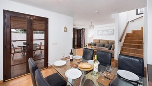 A dining area with a glass table, chairs, and a bottle of wine. There is a sofa and a staircase in the background.