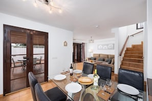 A dining area with a glass table, chairs, and a bottle of wine. There is a sofa and a staircase in the background.
