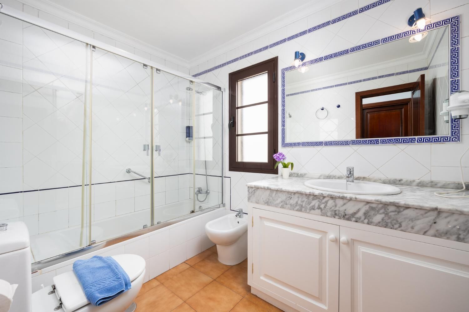 A modern bathroom with a glass-enclosed shower, a white toilet, a sink with a marble countertop, and a large mirror with a decorative frame.
