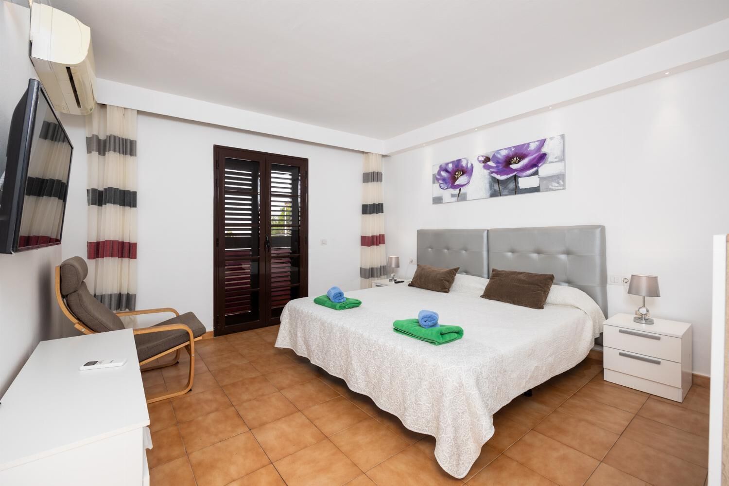 A hotel room with a bed, a chair, a desk, a nightstand, and a wall-mounted TV.