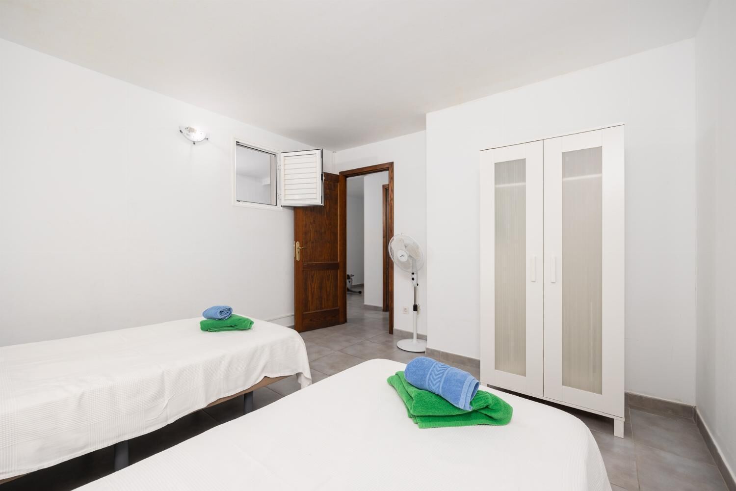 A bedroom with two single beds, a wardrobe, a fan, and a door leading to another room.