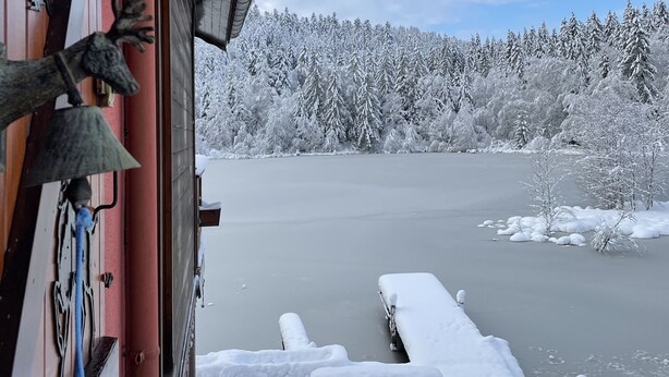 Une vue par la fenêtre sur un paysage enneigé, avec un lac gelé et des arbres recouverts de neige.