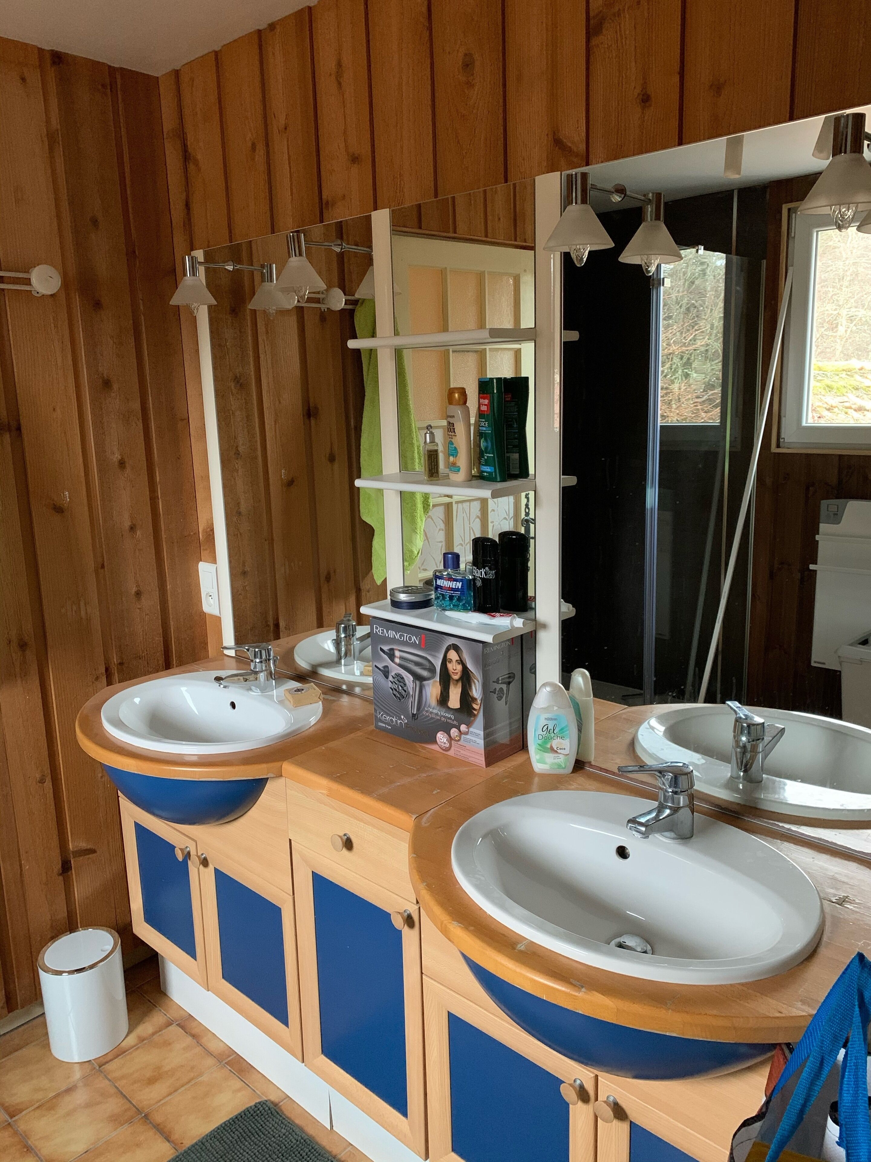 Een badkamer met houten wanden, twee wastafels en een spiegel. Er staan toiletartikelen op de toonbank en een handdoek hangt aan de deur.