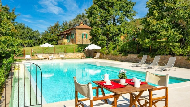 Un área de piscina con una mesa dispuesta para dos, sillas de descanso y un edificio de piedra al fondo.