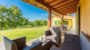 Un patio exterior con mobiliario de mimbre y una mesa, con vistas a un césped cuidado y un edificio amarillo con contraventanas verdes.