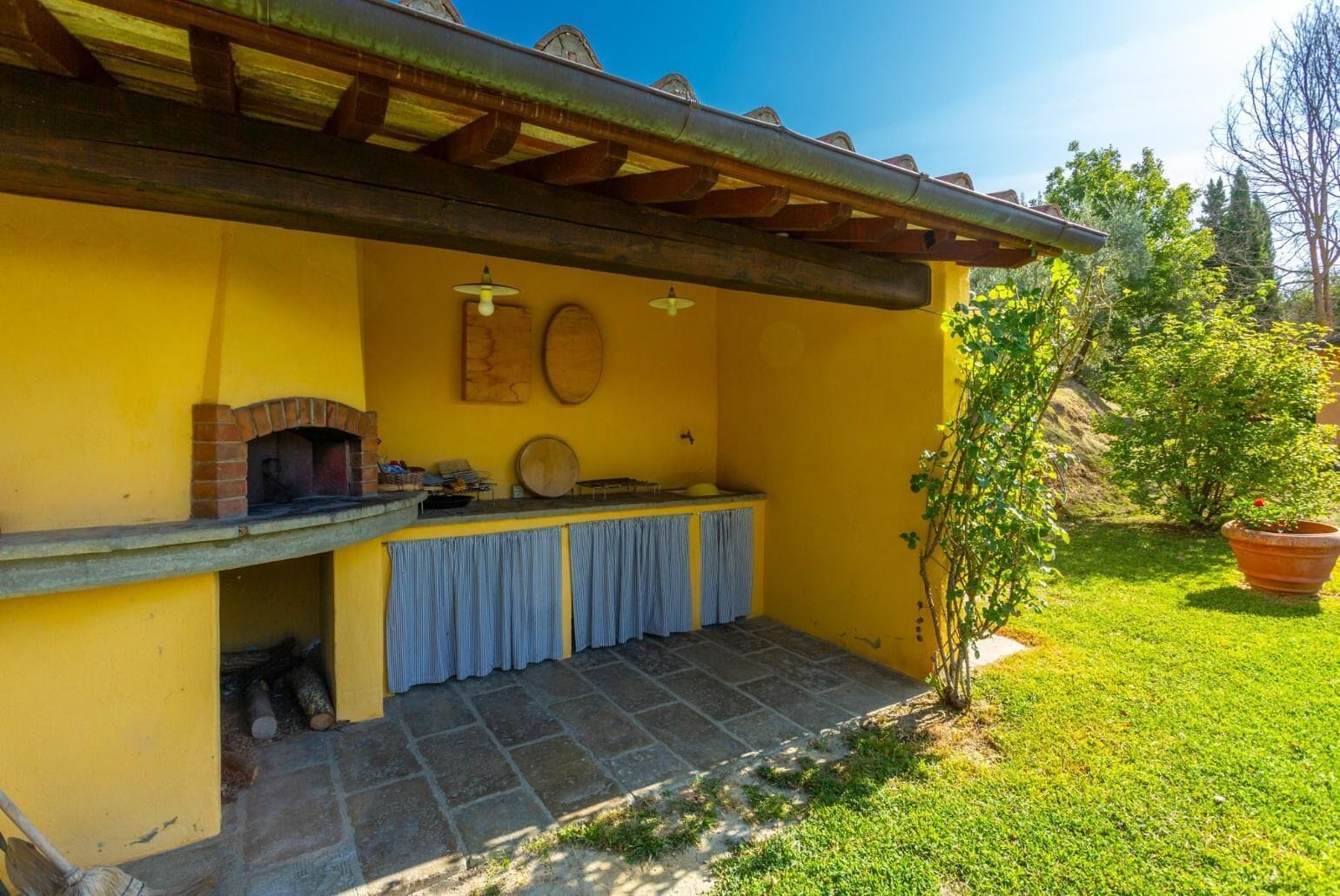 A patio with a brick oven, a wooden table, and a yellow wall.