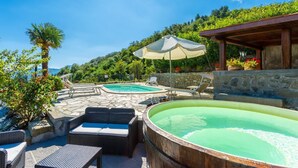 Zona de piscina con sillones de descanso y bañera de hidromasaje, rodeada de suelo de piedra y vegetación.
