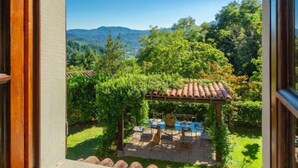 Vista a través de una ventana de un patio con una mesa y sillas, rodeado de follaje y una montaña al fondo.