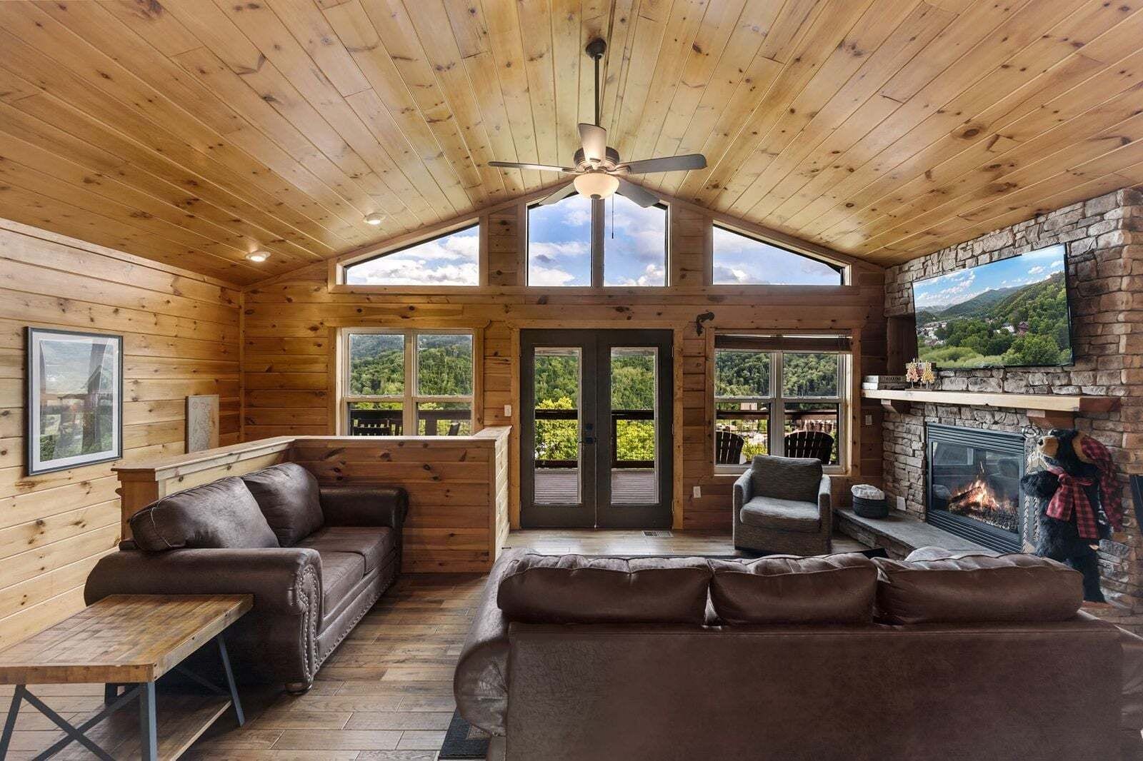 A log cabin interior with a fireplace, leather sofas, a ceiling fan, and large windows offering a scenic view.