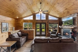 A log cabin interior with a fireplace, leather sofas, a ceiling fan, and large windows offering a scenic view.