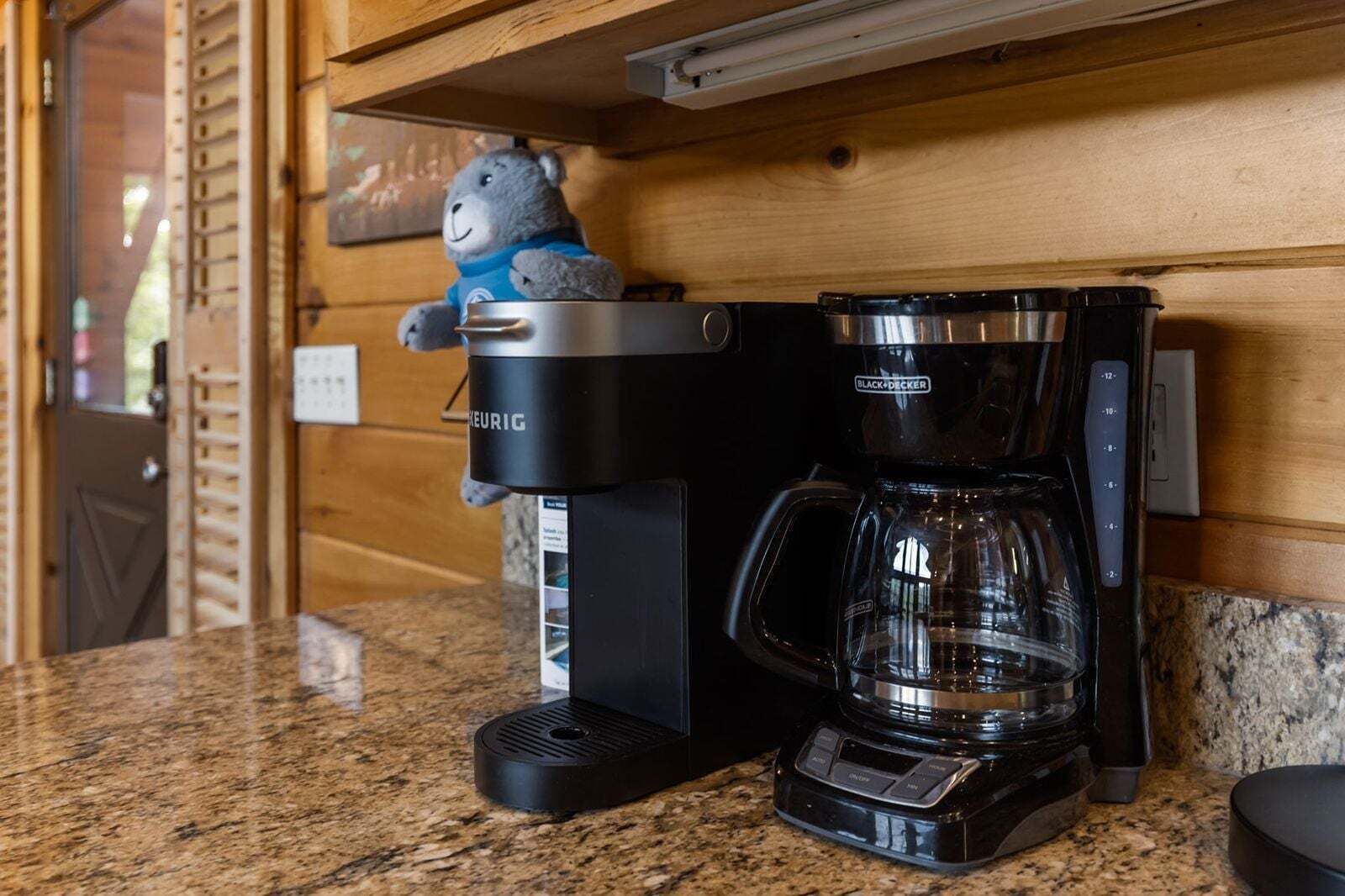 A coffee maker and a coffee pot on a granite countertop in a kitchen with wooden walls.