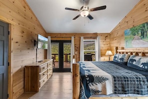 A wooden cabin bedroom with a bed, dresser, TV, and a view of the outdoors.