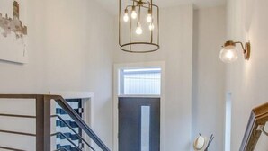 Un escalier avec des marches en bois et une rampe en métal mène à une porte surmontée d’une fenêtre. Un luminaire moderne est suspendu au plafond.