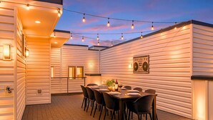 Une terrasse extérieure avec une table à manger pour quatre personnes, entourée de chaises, sous des guirlandes lumineuses.