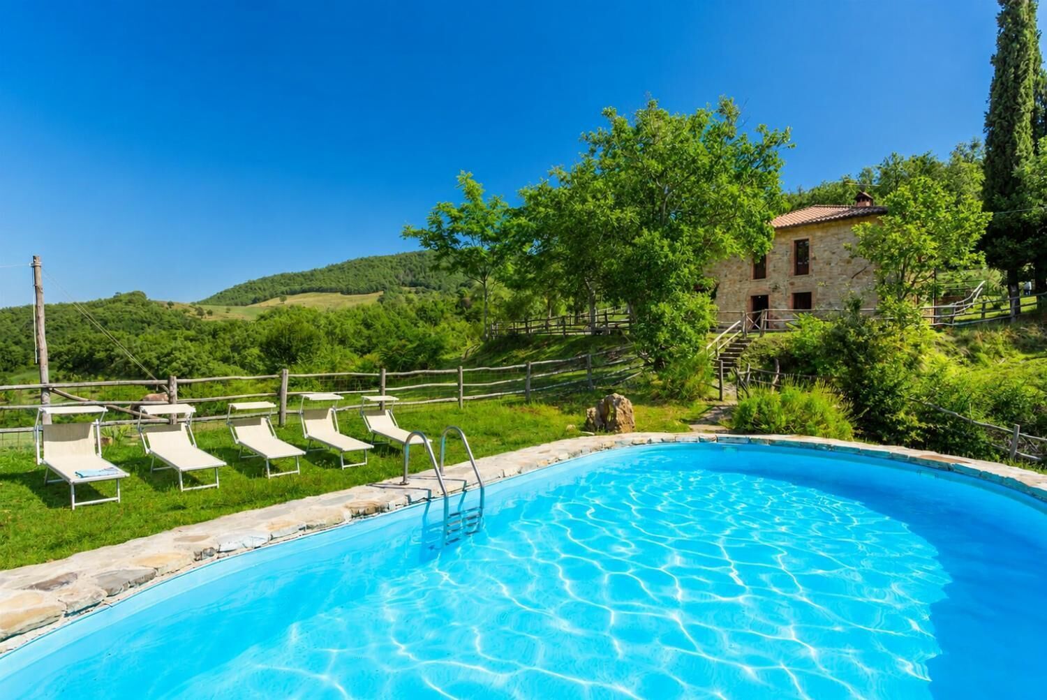 A swimming pool with lounge chairs, a stone building, and a forested hillside in the background.