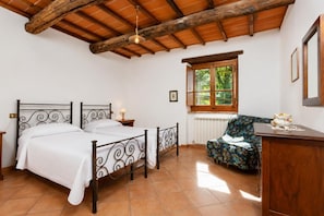 Quarto com teto de madeira, cama de casal, cadeira, radiador e janela com vista para o verde.