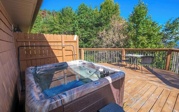 Relax on the deck with a hot tub, table, and chairs, surrounded by trees in a serene natural setting.