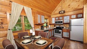 A wooden cabin interior with a dining table set for four, kitchen appliances, and a window with curtains. - Relax Year-Round in Your Private Screened-In Hot Tub Retreat - A Secret Romance (Sevierville)