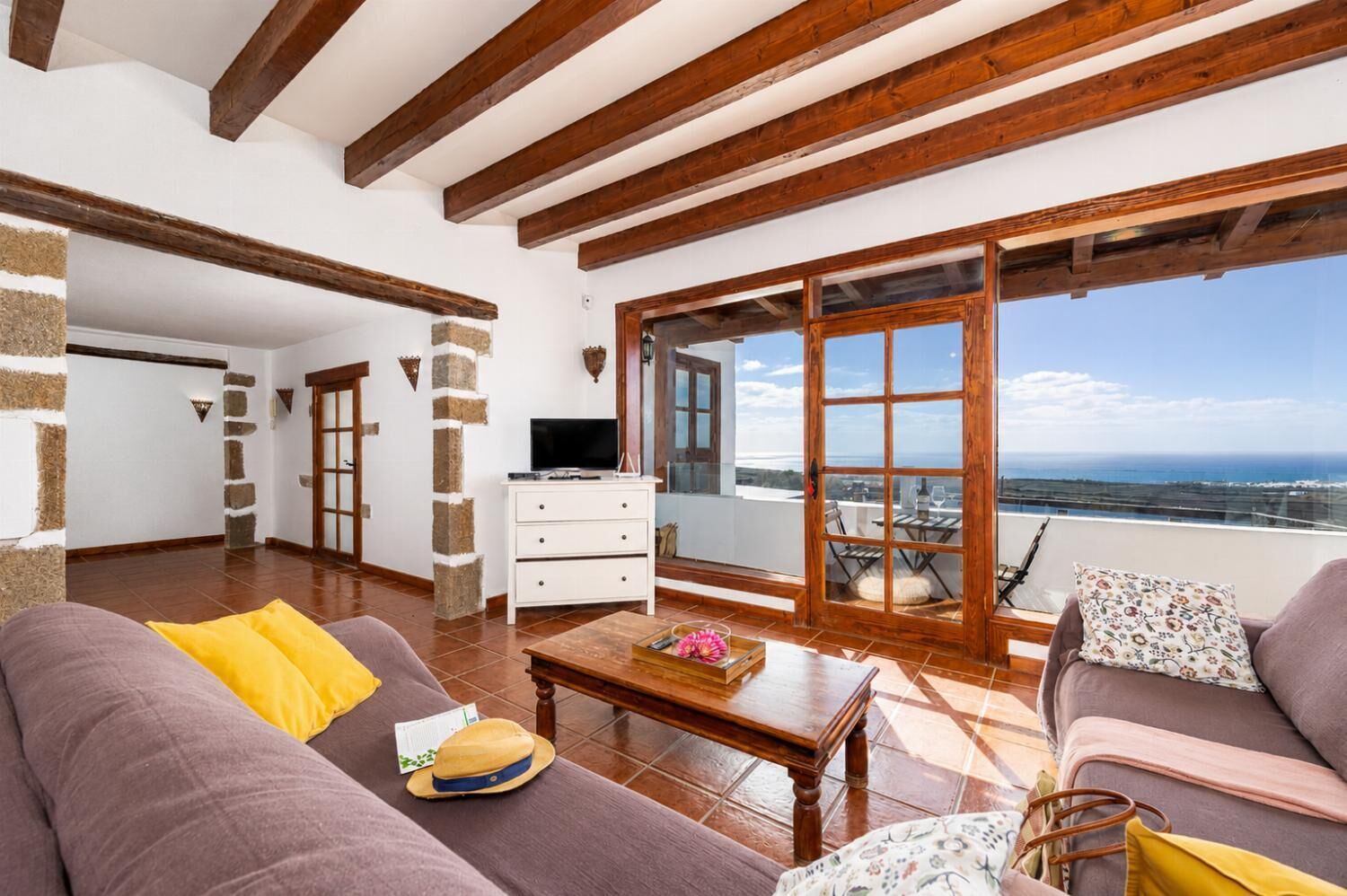 Uma sala de estar ampla com sofá, mesa de centro e televisão. É visível uma varanda com vista para o mar.