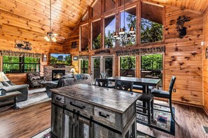 A cabin interior with a fireplace, wooden walls, a dining area, and large windows.