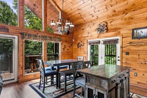 A wooden cabin interior with a dining table, chairs, a chandelier, and a large window. - Amazing Grace - Hot Tub, Stone Firepit & Arcade Loft Near Pigeon Forge (Sevierville)