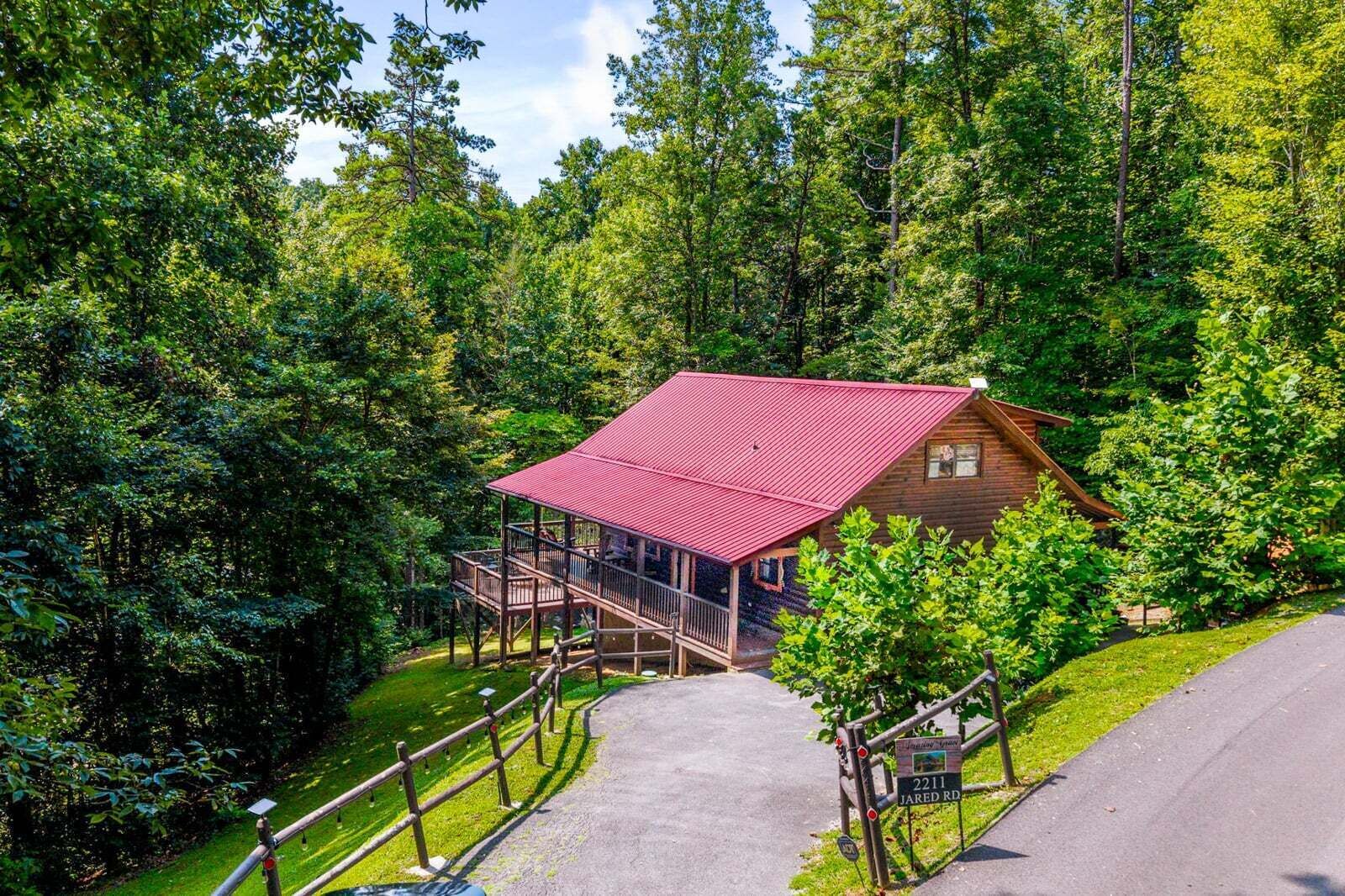 A cabin with a red roof, surrounded by dense forest, accessible by a paved path.