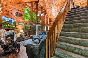 A log cabin interior with a fireplace, a television, a sofa, and a staircase. - Amazing Grace - Hot Tub, Stone Firepit & Arcade Loft Near Pigeon Forge (Sevierville)