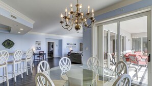Un vaste espace dédié à la salle à manger, avec une table à plateau de verre entourée de chaises blanches, un lustre et une vue sur un espace extérieur aménagé, accessible par de grandes portes-fenêtres coulissantes.