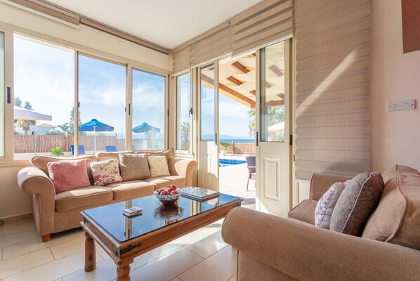 Un salon chaleureux avec un canapé beige, une table basse en verre et une vue sur un jardin avec une piscine et des parasols.
