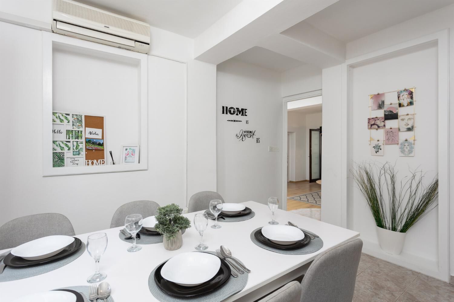 Une salle à manger moderne avec une table blanche dressée pour quatre personnes, des chaises grises et une petite plante servant de centre de table.