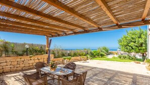 Eine überdachte Terrasse mit Korbmöbeln, einer Steinwand und Blick auf das Meer.