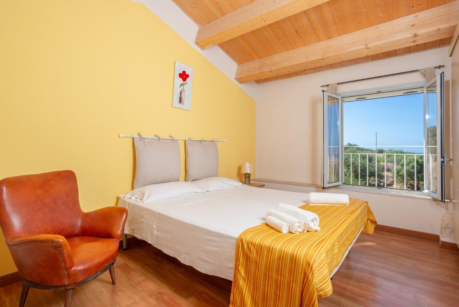 A hotel room with a bed, a chair, a window with a view, and a painting on the wall.