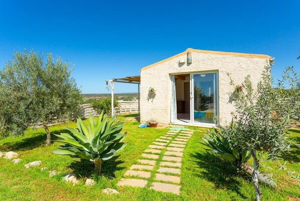 A small, single-story building with a stone exterior and a glass door, surrounded by a well-maintained garden with olive trees and a stone pathway.